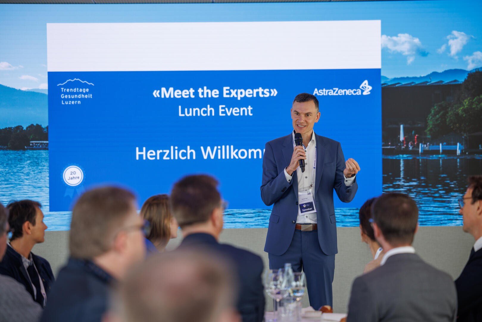 Ein Mann in einem blauen Anzug spricht in ein Mikrofon bei einer "Meet the Experts"-Lunch-Veranstaltung von AstraZeneca, während die Teilnehmer sitzen und auf einem Bildschirm hinter ihm "Herzlich Willkommen" und Bilder eines Sees zu sehen sind.