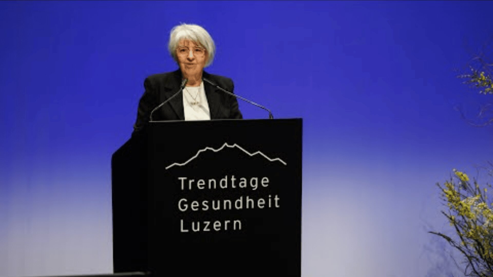 Eine ältere Frau mit kurzen grauen Haaren steht an einem Podium mit der Aufschrift Trendtage Gesundheit Luzern vor einem blauen Hintergrund und spricht an einer Veranstaltung oder Konferenz.