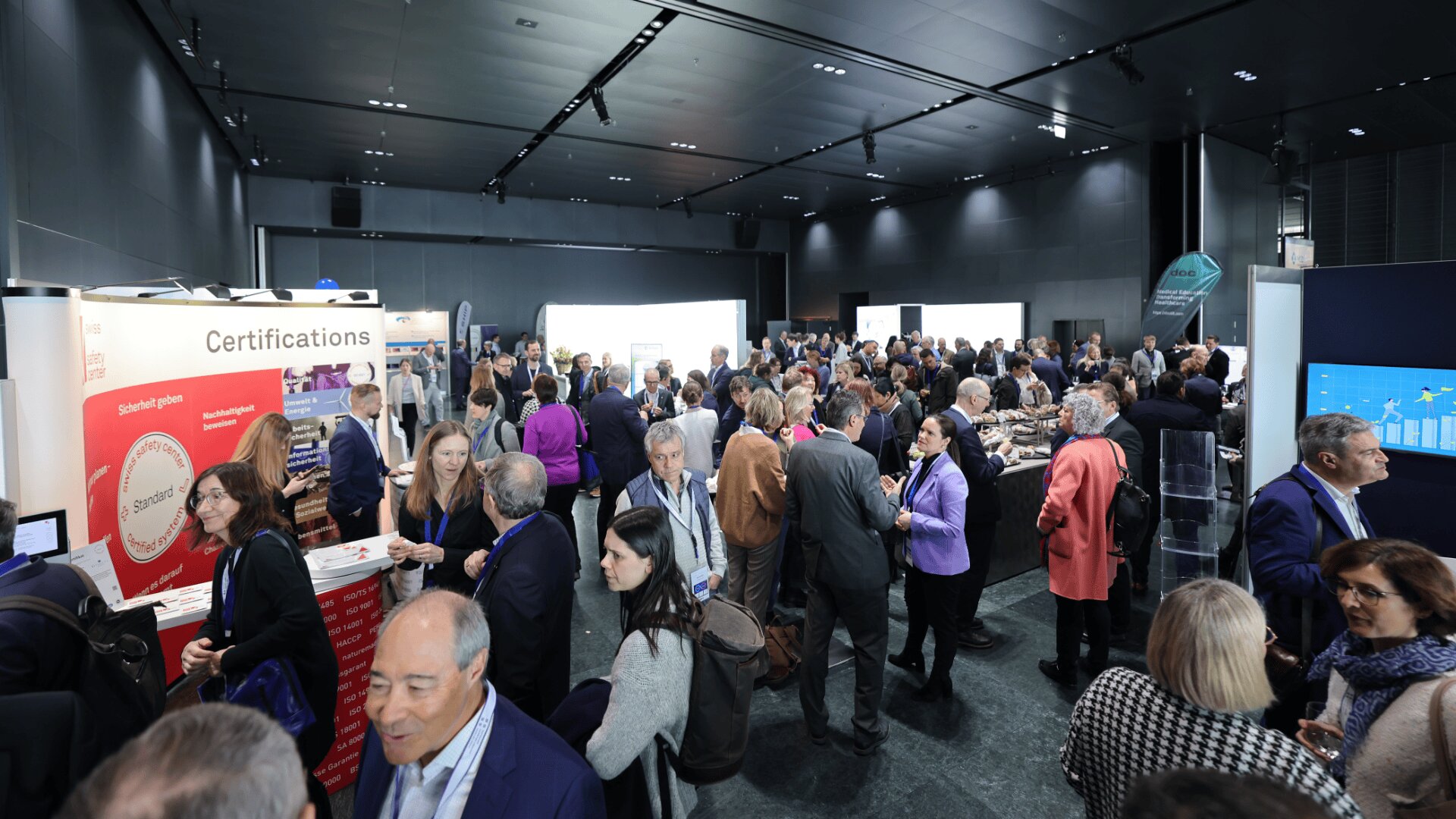 Eine große Gruppe von Menschen trifft sich auf einer Fachkonferenz oder Ausstellung und knüpft Kontakte. Stände und Displays zu Zertifizierungen sind sichtbar, und die Teilnehmer sind in einem modernen, gut beleuchteten Veranstaltungsort in Gespräche vertieft.