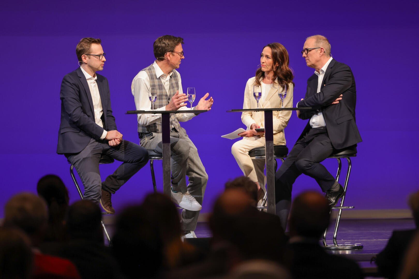 Vier Personen in Geschäftskleidung sitzen auf hohen Stühlen um zwei Tische herum und führen eine Podiumsdiskussion auf der Bühne vor einem violetten Hintergrund, während im Vordergrund Wassergläser und das Publikum zu sehen sind.