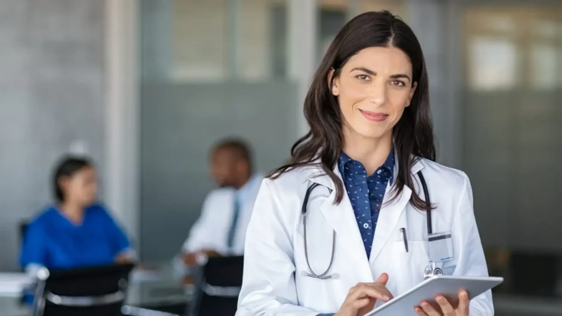 Eine Ärztin mit dunklem Haar, weißem Kittel und Stethoskop, lächelt, während sie ein Tablet benutzt. Im unscharfen Hintergrund sitzen zwei Personen an einem Tisch und führen ein Gespräch.