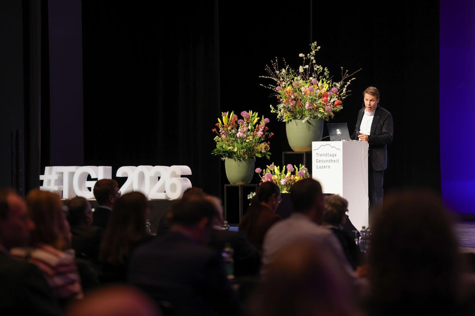 Ein Mann im Anzug spricht an einem Podium mit Blumengestecken und einem Schild mit der Aufschrift Trendtage Gesundheit Luzern. In grossen weissen Buchstaben steht im Hintergrund #TGL2026, während das Publikum zuhört.