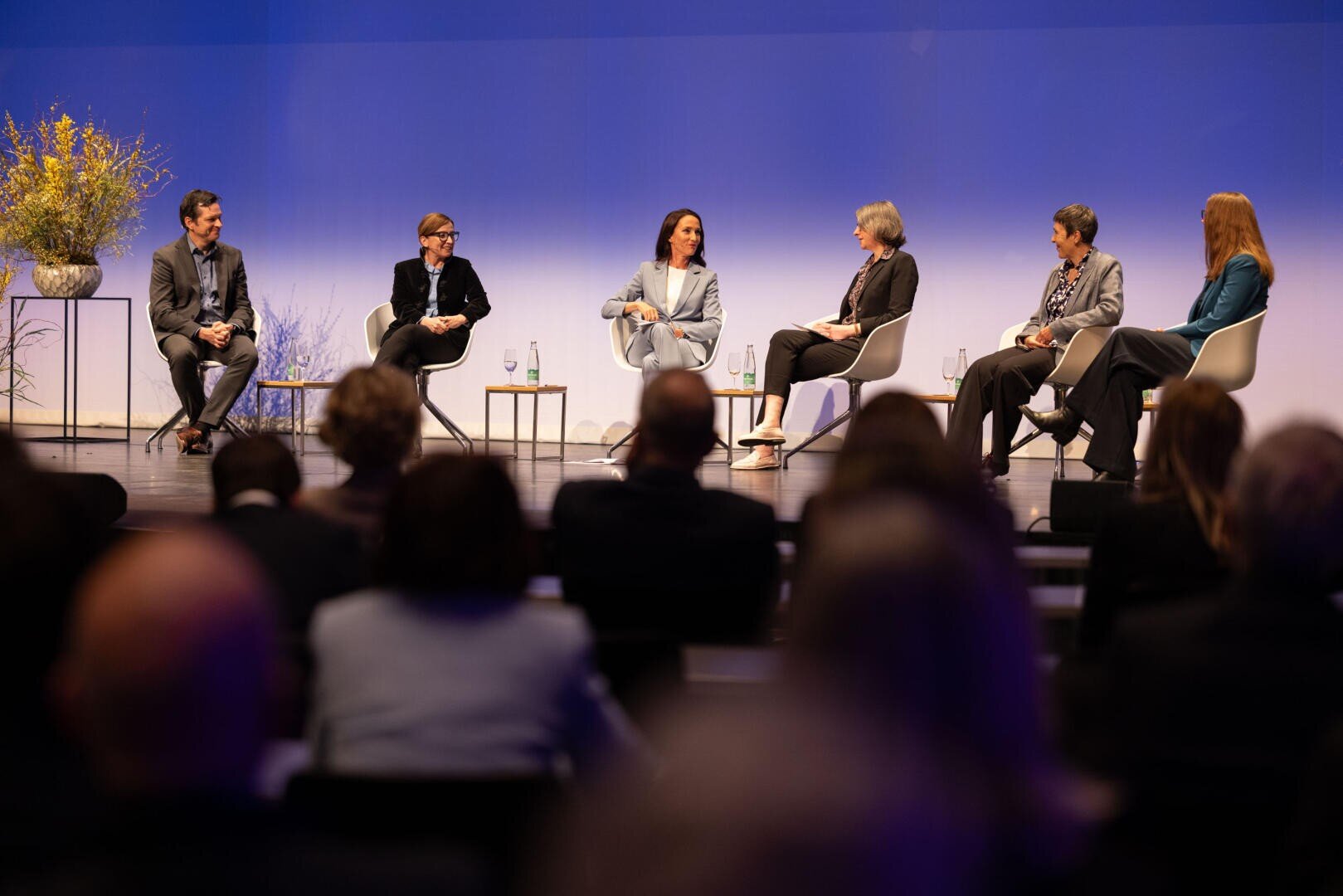 Fünf Podiumsteilnehmer sitzen auf der Bühne und führen eine Diskussion, während das Publikum zusieht. Der Hintergrund hat einen blauen Farbverlauf, und auf der Bühne sind gelbe Zierblumen zu sehen. Die Diskussionsteilnehmer sind in Geschäftskleidung gekleidet.
