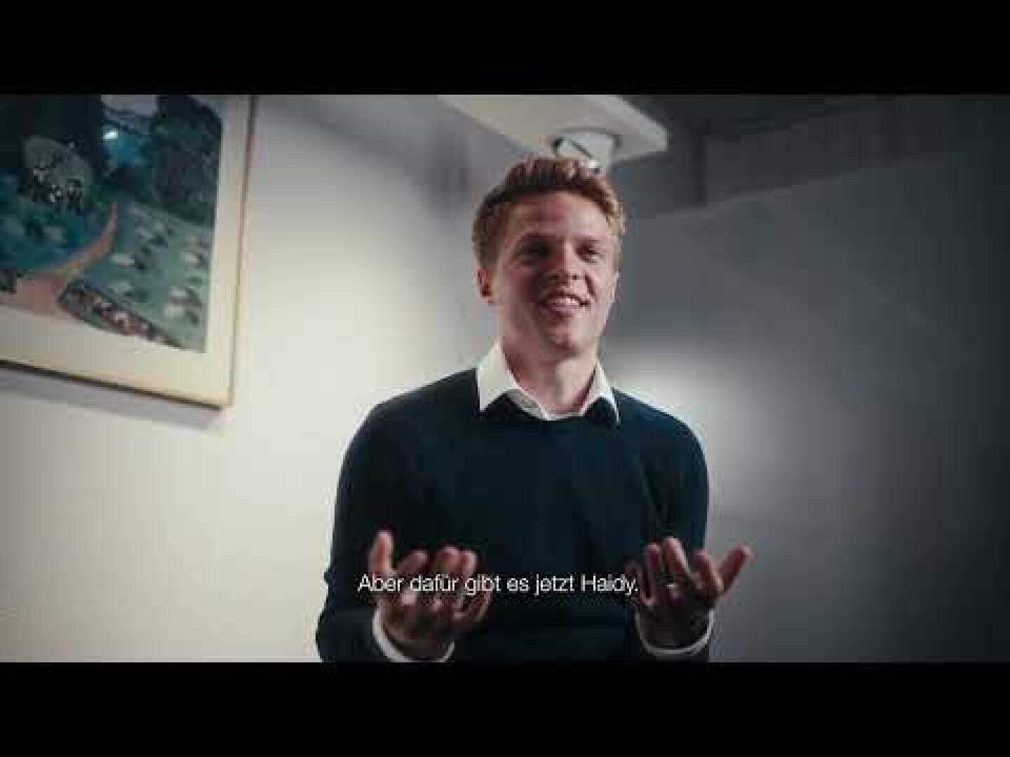 Ein junger Mann mit blondem Haar, der einen dunklen Pullover über einem weißen Hemd trägt, sitzt in einem Haus und lächelt und gestikuliert mit den Händen. Am unteren Rand des Bildes sind deutsche Untertitel zu sehen. Hinter ihm hängt ein gerahmtes Bild an der Wand.