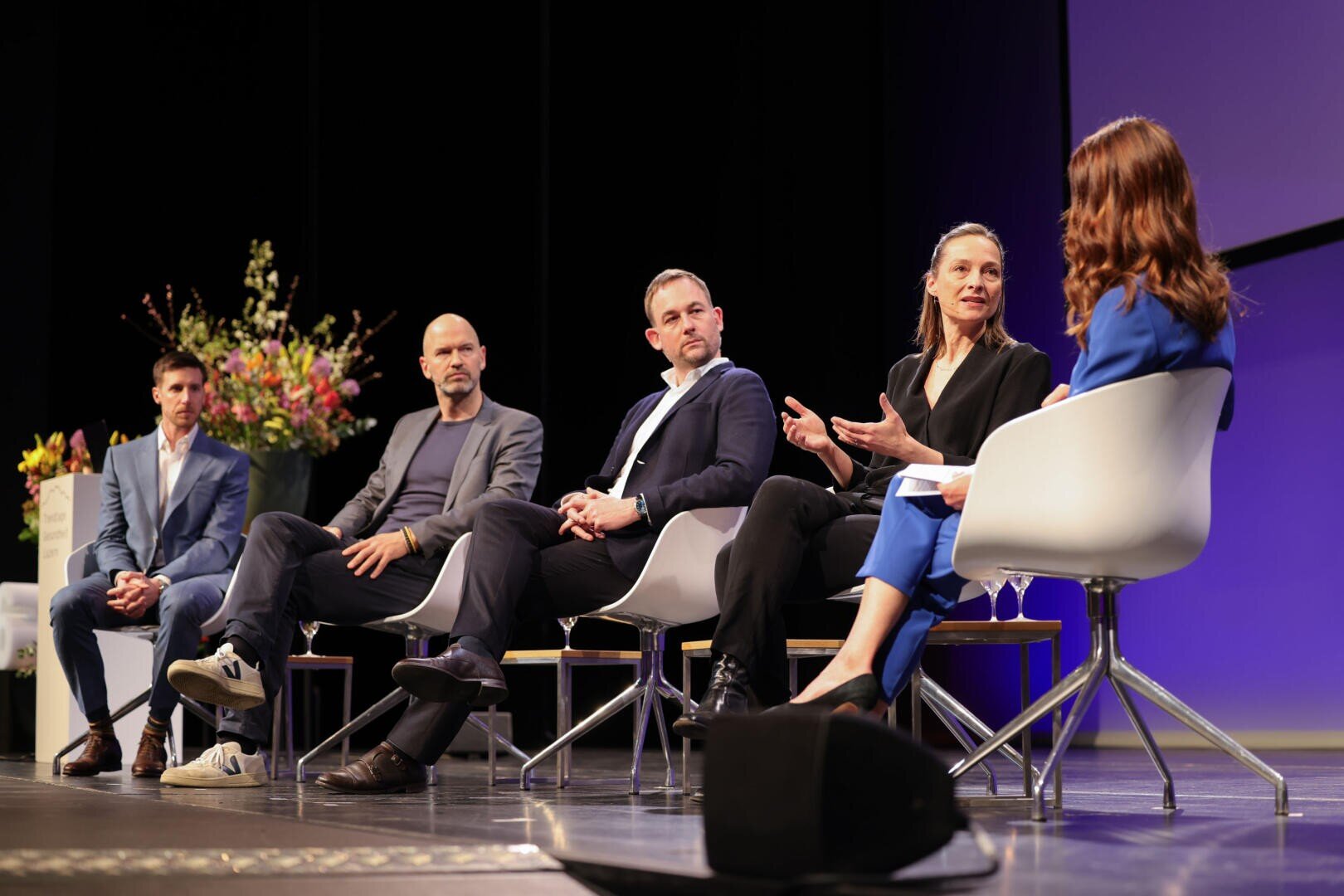 Fünf Personen in Geschäftskleidung sitzen auf einer Bühne in modernen Stühlen und führen eine Podiumsdiskussion. Eine Frau gestikuliert, während sie spricht. Im Hintergrund sind Blumen und ein großer Bildschirm mit violetter Beleuchtung zu sehen.