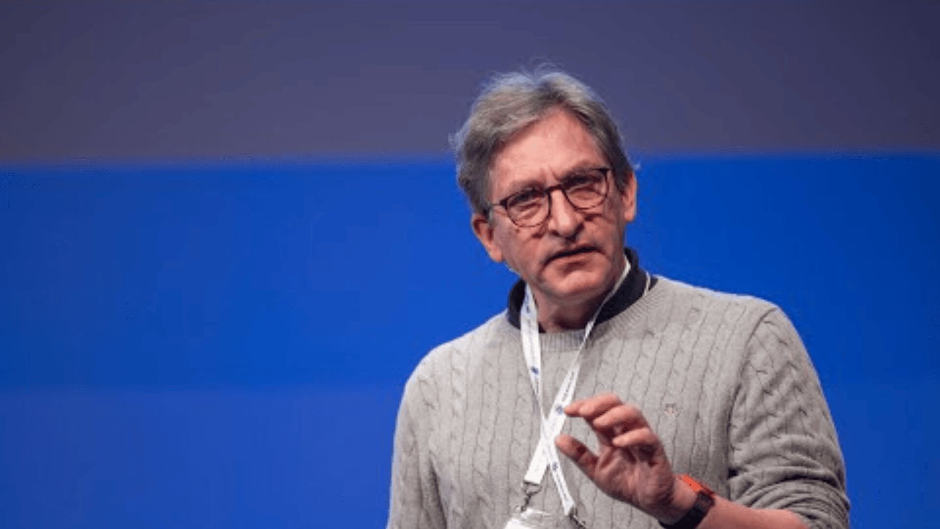 Ein Mann mit grauem Haar und Brille, der einen grauen Strickpullover und ein weißes Schlüsselband trägt, gestikuliert, während er auf der Bühne vor einem blauen Hintergrund spricht.