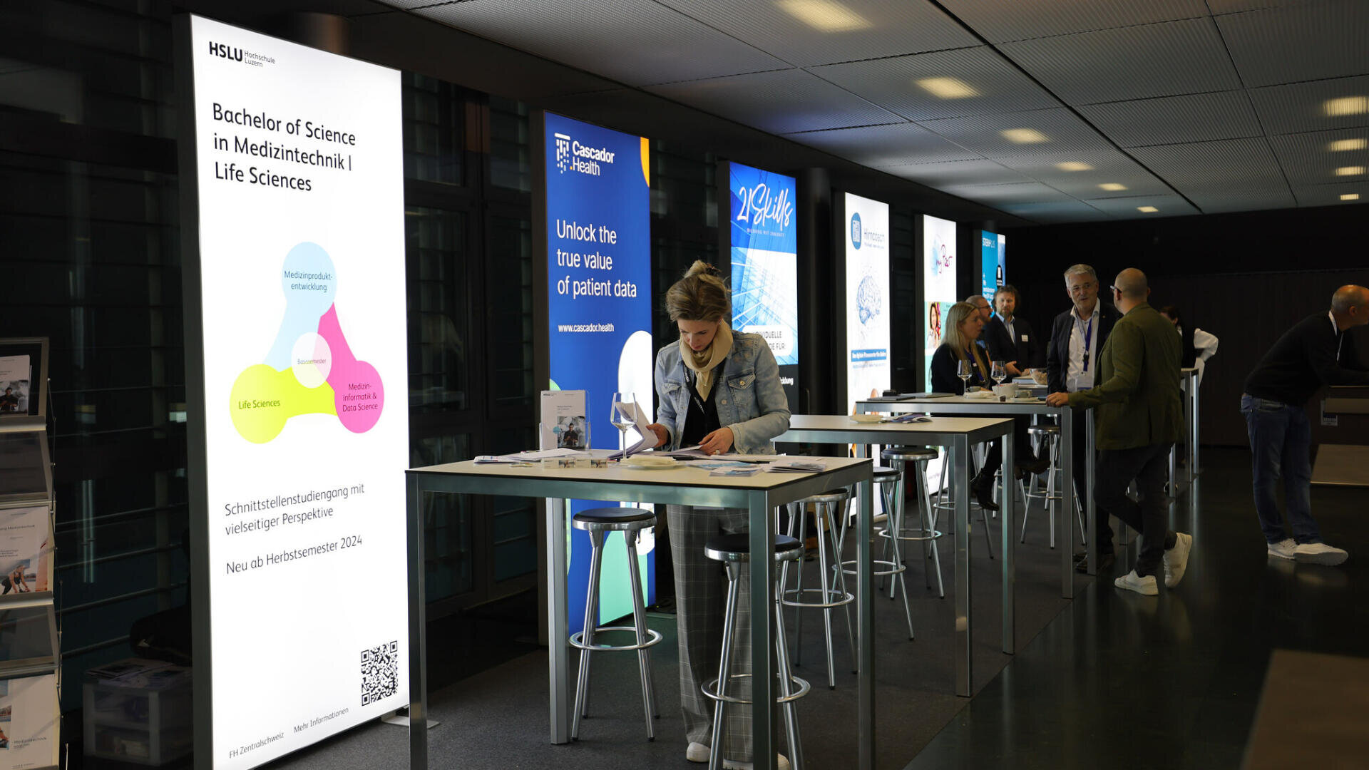 An einer Reihe von Informationsständen mit hohen Tischen und Hockern tauschen sich die Besucher aus. Große beleuchtete Schilder zeigen Bildungs- und Gesundheitsbotschaften, darunter Bachelor of Science in Medizintechnik | Life Sciences.