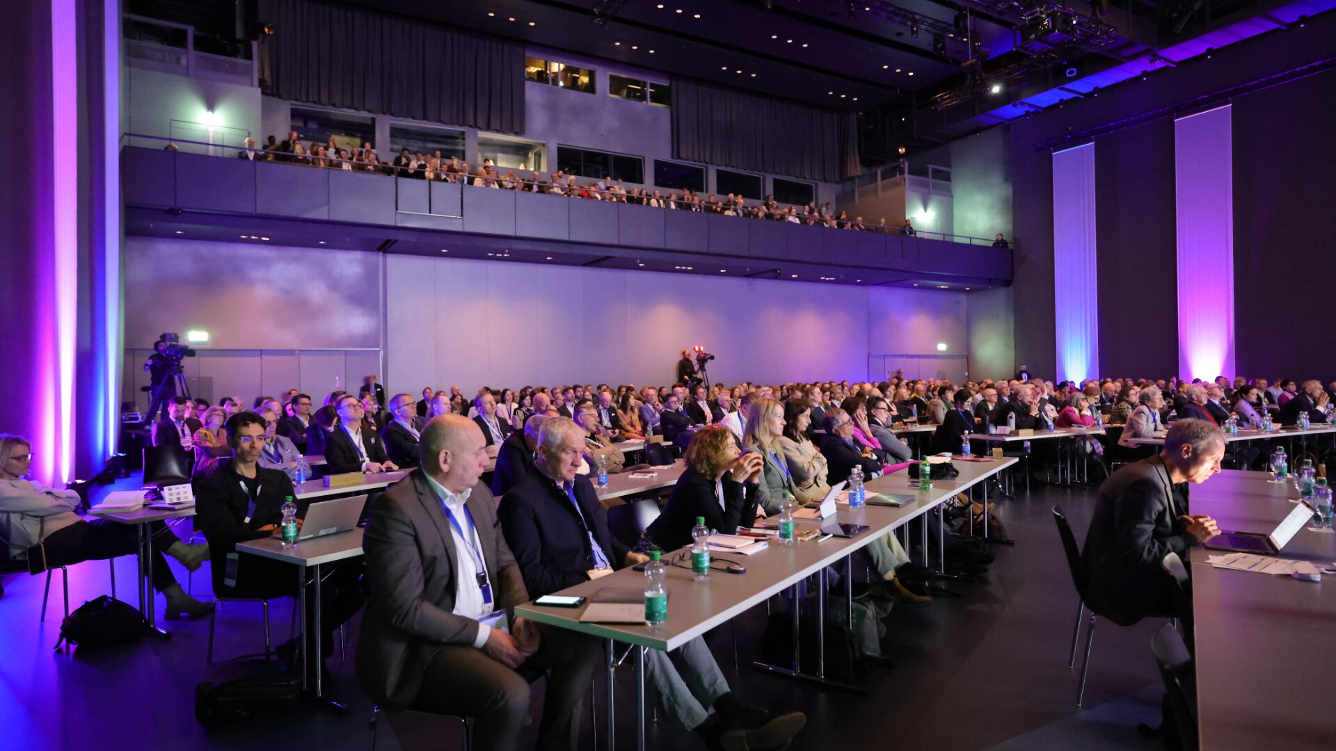 Ein großes Publikum sitzt an langen Tischen in einem modernen Konferenzsaal und verfolgt aufmerksam eine Präsentation. Der Raum ist schwach beleuchtet, mit violetter Akzentbeleuchtung, und auch auf einem oberen Balkon sitzen einige Personen.