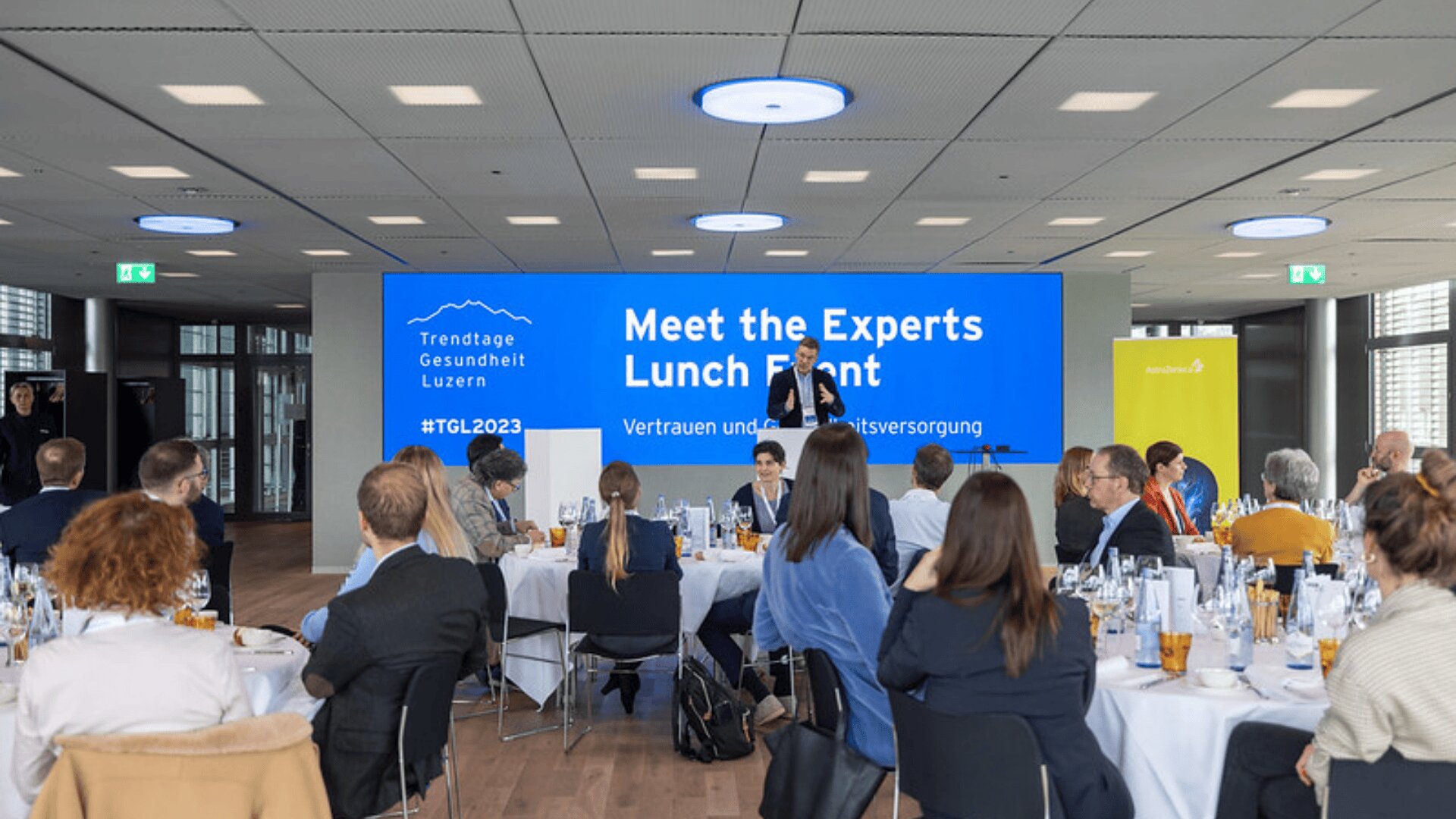 An runden Tischen sitzende Personen während einer professionellen Mittagsveranstaltung mit Blick auf einen großen Bildschirm mit der Aufschrift Meet the Experts Lunch Event in einem modernen, gut beleuchteten Konferenzraum.