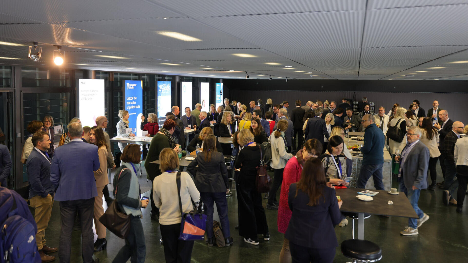 Eine große Gruppe von Menschen knüpft Kontakte in einem modernen, überdachten Konferenzraum. Sie stehen um Stehtische herum und unterhalten sich, während im Hintergrund Informationstafeln zu sehen sind.