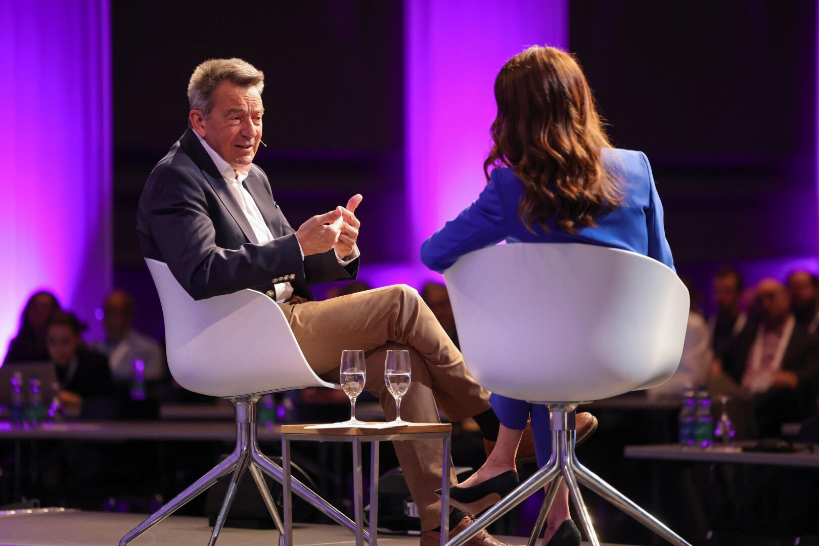 Ein Mann und eine Frau sitzen in modernen weißen Stühlen auf der Bühne und führen ein Gespräch. Beide haben Mikrofone. Der Hintergrund ist mit violetten Lichtern beleuchtet, und in dem schwach beleuchteten Raum befindet sich ein Publikum. Auf einem kleinen Tisch stehen zwei Gläser mit Wasser.