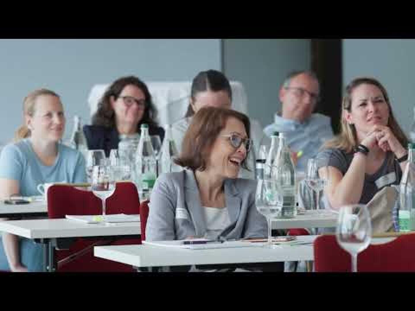 Eine Gruppe von Erwachsenen sitzt an Tischen in einem Klassenzimmer oder Konferenzraum, einige lächeln und lachen. Auf den Tischen stehen Wasserflaschen und Gläser, und der Fokus liegt auf einer Frau im Vordergrund, die lächelt.