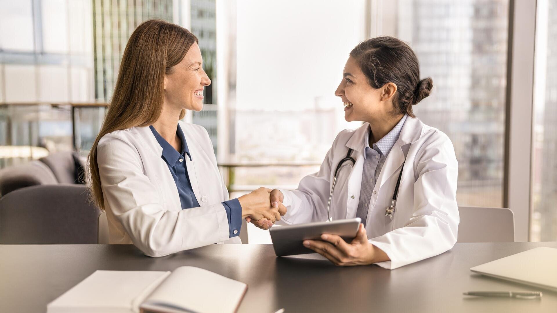Zwei Frauen in weißen Kitteln, eine mit einem Stethoskop, sitzen an einem Schreibtisch in einem Büro, lächeln und schütteln sich die Hände. Auf dem Schreibtisch liegen ein aufgeschlagenes Notebook und ein Tablet, und große Fenster zeigen städtische Gebäude im Hintergrund.