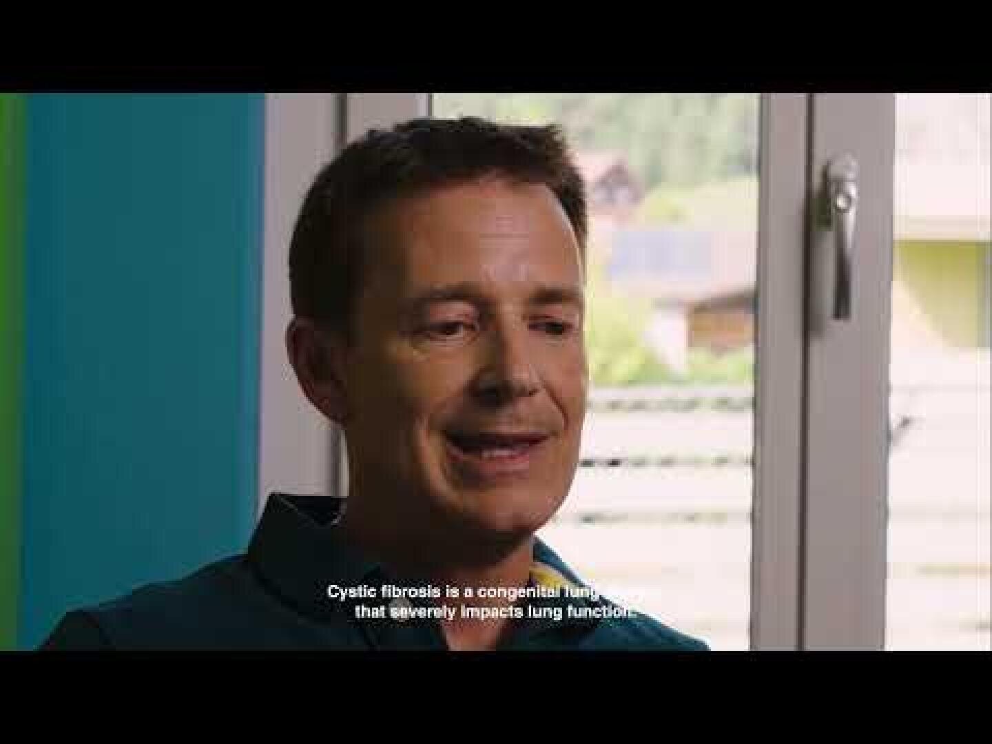 Ein Mann mit kurzen braunen Haaren sitzt in einem Haus in der Nähe eines Fensters. Er spricht, und die Untertitel lauten: Mukoviszidose ist eine angeborene Krankheit, die die Lungenfunktion stark beeinträchtigt.