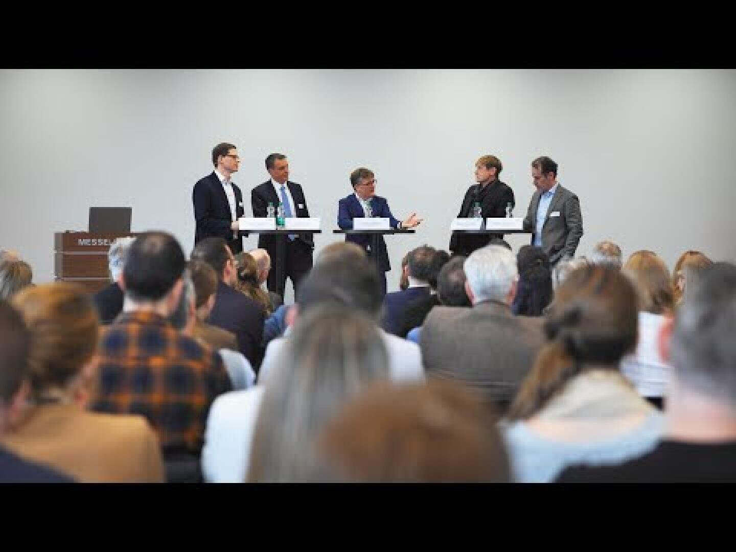 Ein Gremium von fünf Personen in Anzügen sitzt an einem Tisch auf einer Bühne und diskutiert mit Blick auf das Publikum in einer Konferenz- oder Seminarumgebung. Die Zuhörer sitzen und hören aufmerksam zu.