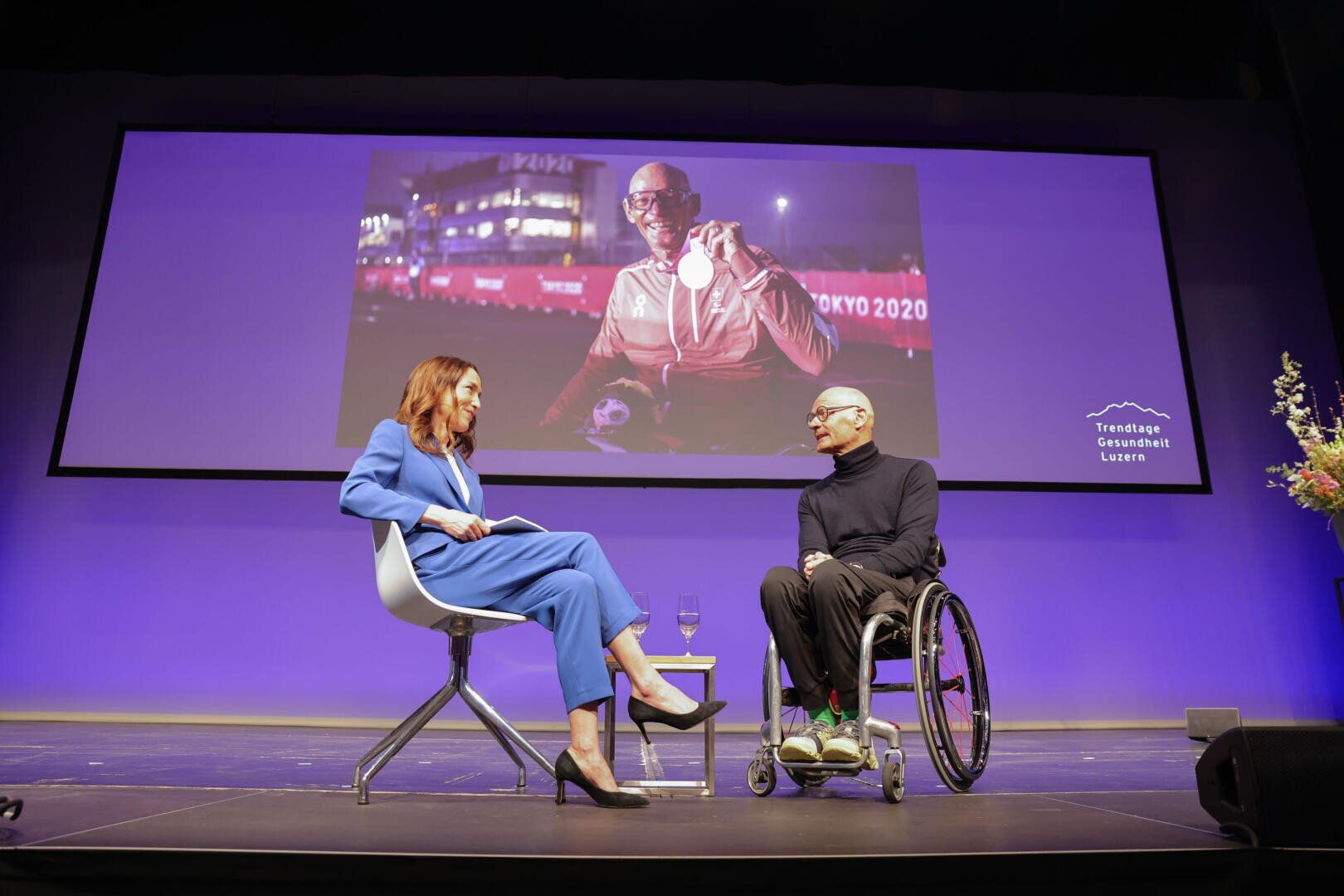Eine Frau in einem blauen Anzug interviewt einen Mann im Rollstuhl auf der Bühne. Hinter ihnen ist ein großer Bildschirm zu sehen, der einen Sportler mit einer Medaille von Tokio 2020 zeigt. Die Szenerie ist hell beleuchtet, mit einem lila Hintergrund und Blumen auf der rechten Seite.