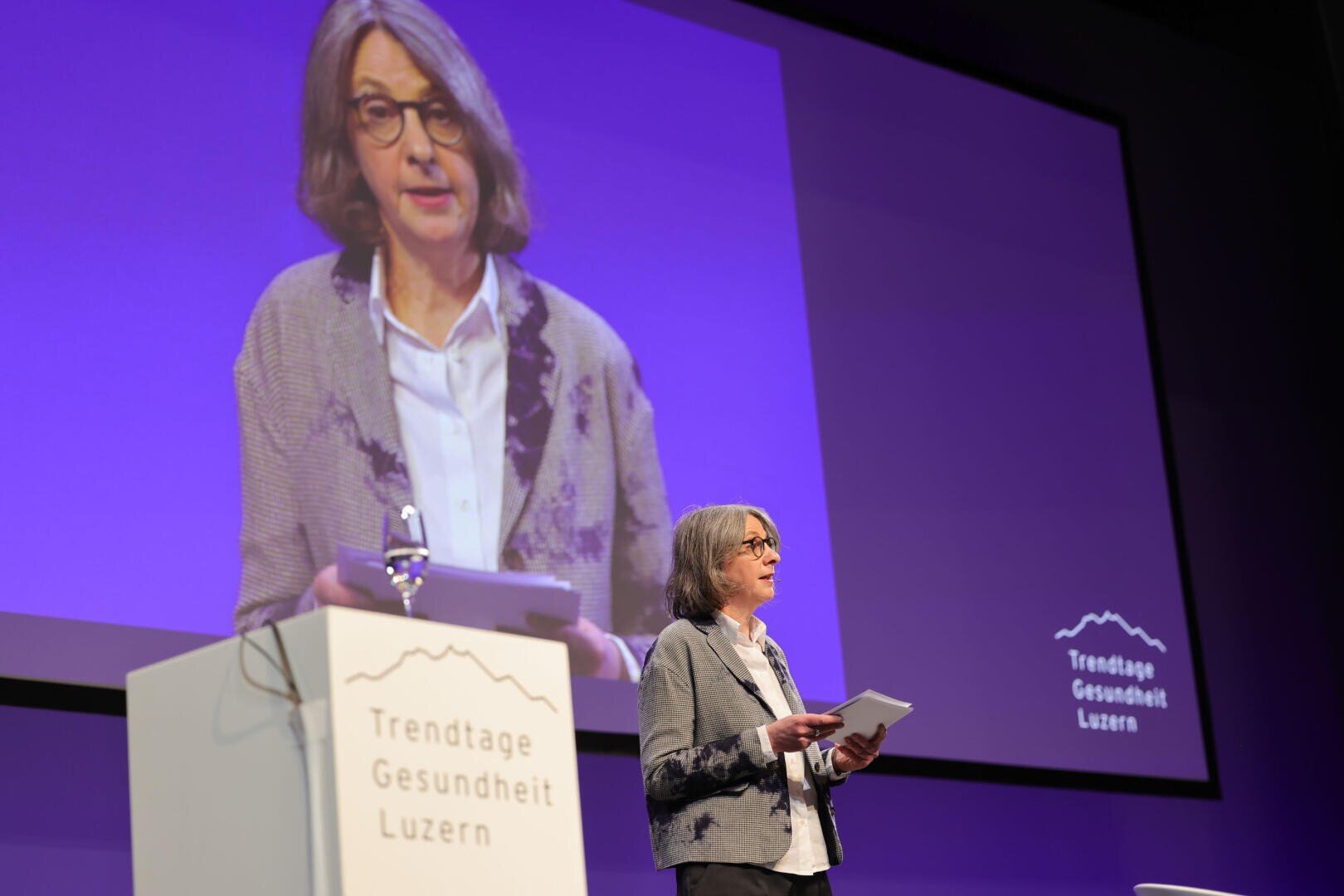 Eine Frau mit einem Tablet in der Hand spricht auf der Bühne der Trendtage Gesundheit Luzern, während ihr Bild auf eine grosse Leinwand hinter ihr projiziert wird. Sie steht neben einem Podium, auf dem der Name der Veranstaltung steht, vor einem violetten Hintergrund.