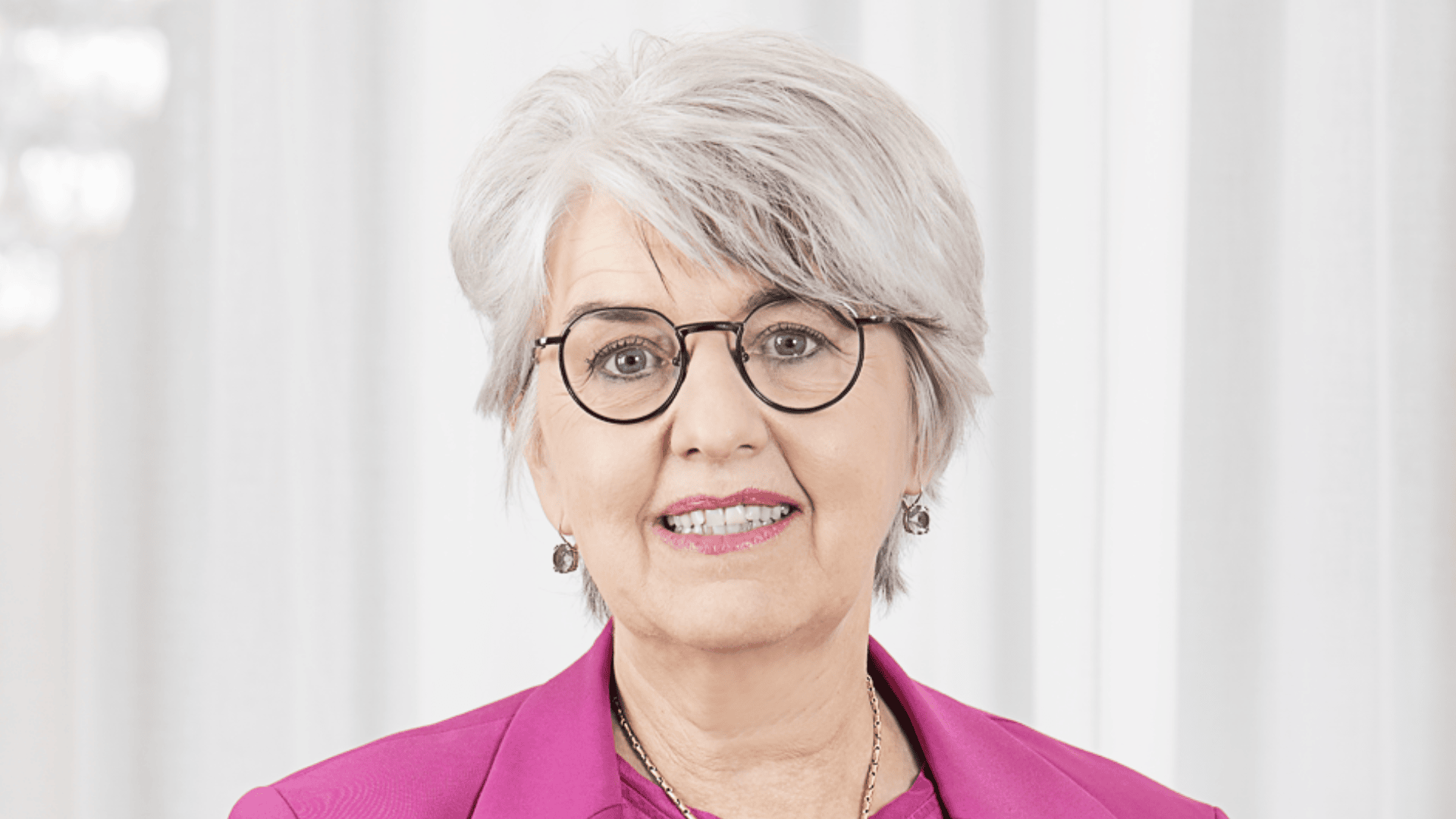 Eine lächelnde ältere Frau mit kurzen grauen Haaren, die eine runde Brille, einen rosa Blazer, Ohrringe und eine Halskette trägt, steht vor einem weißen Vorhang im Hintergrund.
