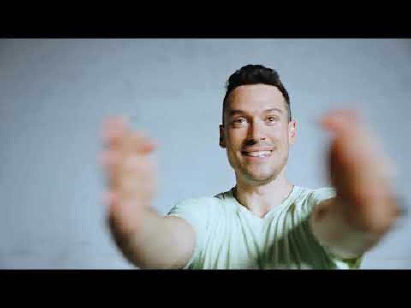 Ein lächelnder Mann in einem hellen Hemd steht vor einem schlichten Hintergrund und streckt beide Arme nach vorne aus, als ob er sich umarmen wollte.
