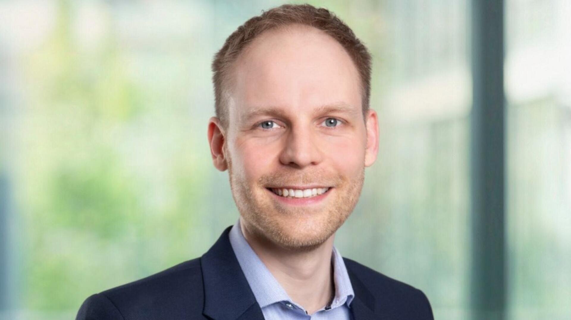 Ein Mann in einem marineblauen Blazer und einem hellblauen Hemd lächelt, während er in einem Innenraum vor großen Fenstern mit einem unscharfen, grünen Hintergrund im Freien steht.
