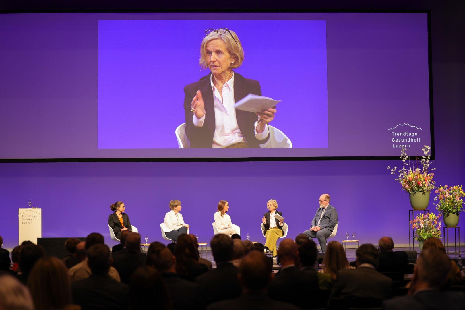 Eine Gruppe von fünf Personen sitzt auf der Bühne und diskutiert über Gesundheitsthemen, wobei eine Frau mit Papieren in der Hand spricht. Ihr Bild wird auf eine große Leinwand hinter ihnen projiziert. Das Publikum hört aufmerksam zu. Blumen schmücken die Bühne.