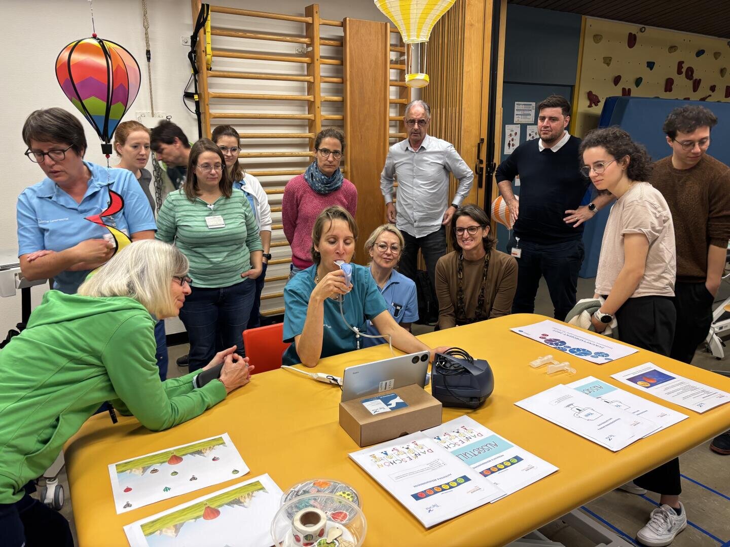 Eine Gruppe Erwachsener sitzt um einen Tisch mit Papieren und einem Tablet und sieht sich eine Vorführung an. Der Raum ist mit Unterrichtsmaterialien, einer Heißluftballon-Dekoration und einer Sprossenwand ausgestattet.