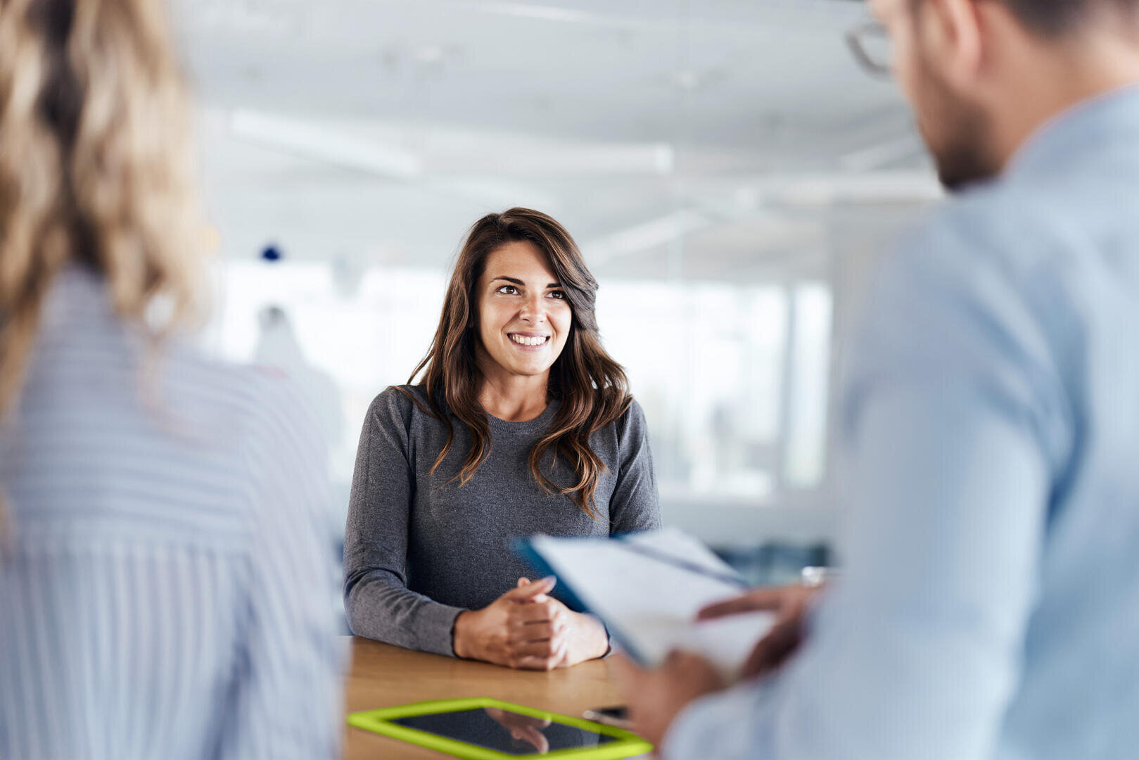 Eine Frau sitzt an einem Tisch, lächelt und spricht mit zwei Personen, die vor ihr stehen. Die eine Person hält ein Klemmbrett. Ein Tablet liegt auf dem Tisch. Der Schauplatz ist ein heller, moderner Büroraum.