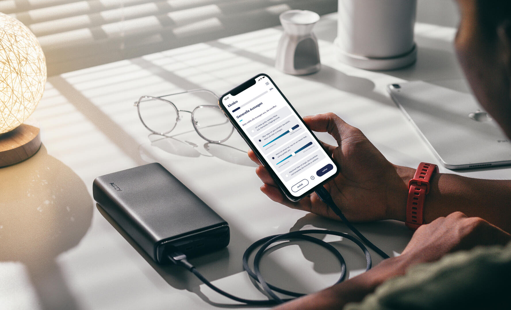 Eine Person, die eine rote Smartwatch trägt, hält ein Smartphone, das an ein tragbares Ladegerät angeschlossen ist, auf einem Schreibtisch mit einer Brille, einer Lampe und einer Tasse, wobei das Sonnenlicht durch die Jalousien fällt.