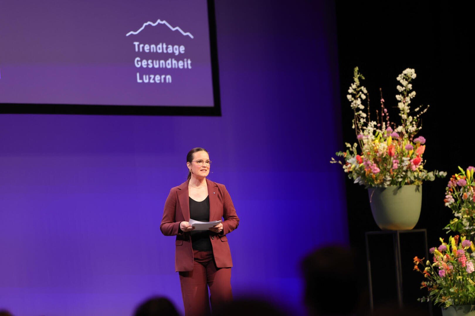 Eine Person in einem kastanienbraunen Anzug steht auf der Bühne und hält Papiere in der Hand, daneben steht ein großes Blumenarrangement. Auf einem projizierten Schild steht "Trendtage Gesundheit Luzern" auf violettem Hintergrund.