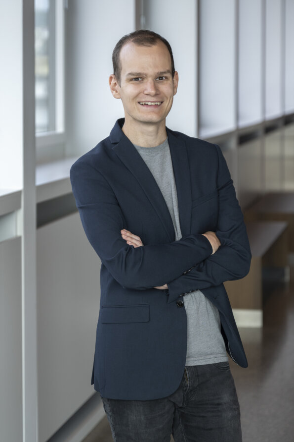 Ein Mann mit kurzen Haaren, einem dunklen Blazer über einem grauen Hemd und schwarzen Jeans, steht mit verschränkten Armen in einem Haus und lächelt in die Kamera. Er steht neben großen Fenstern und einem Flur mit Bänken.